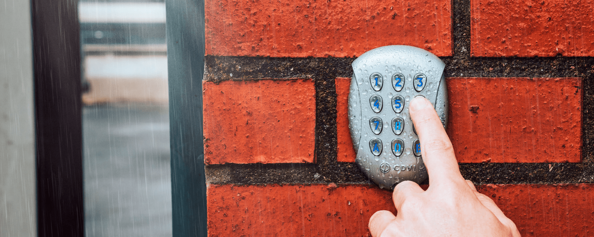 A silver access control keypad is mounted on a bright orange brick wall outdoors in the rain. A person's hand is reaching out to press the keys