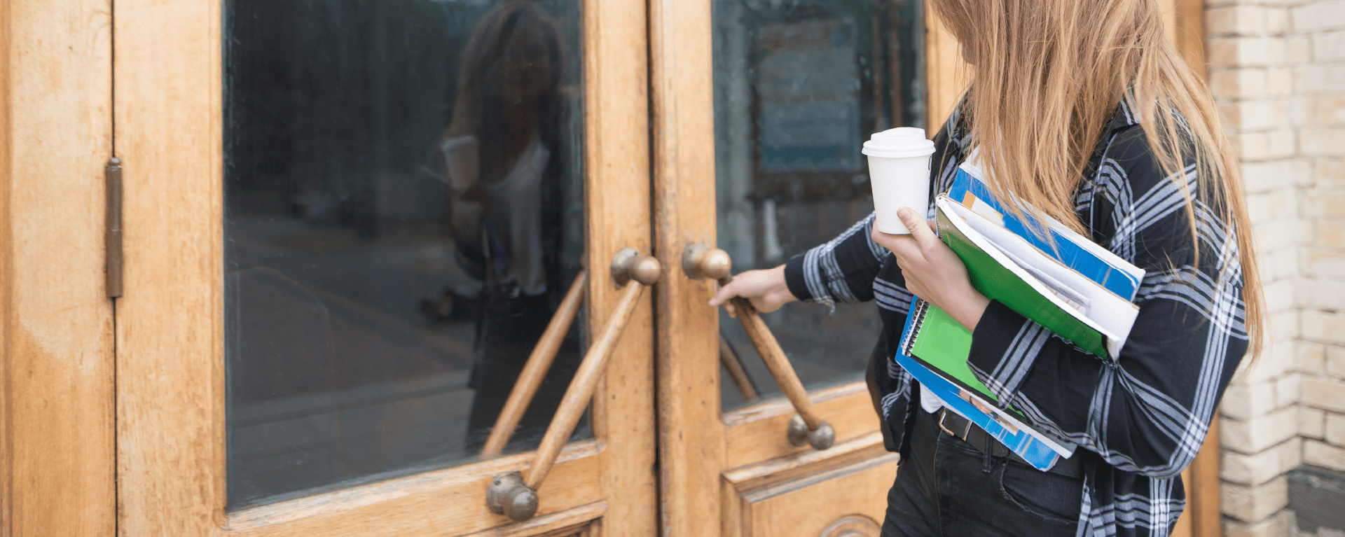 A woman in casual clothes is reaching out to pull a large wooden door open while carrying folders and a takeaway cup of coffee